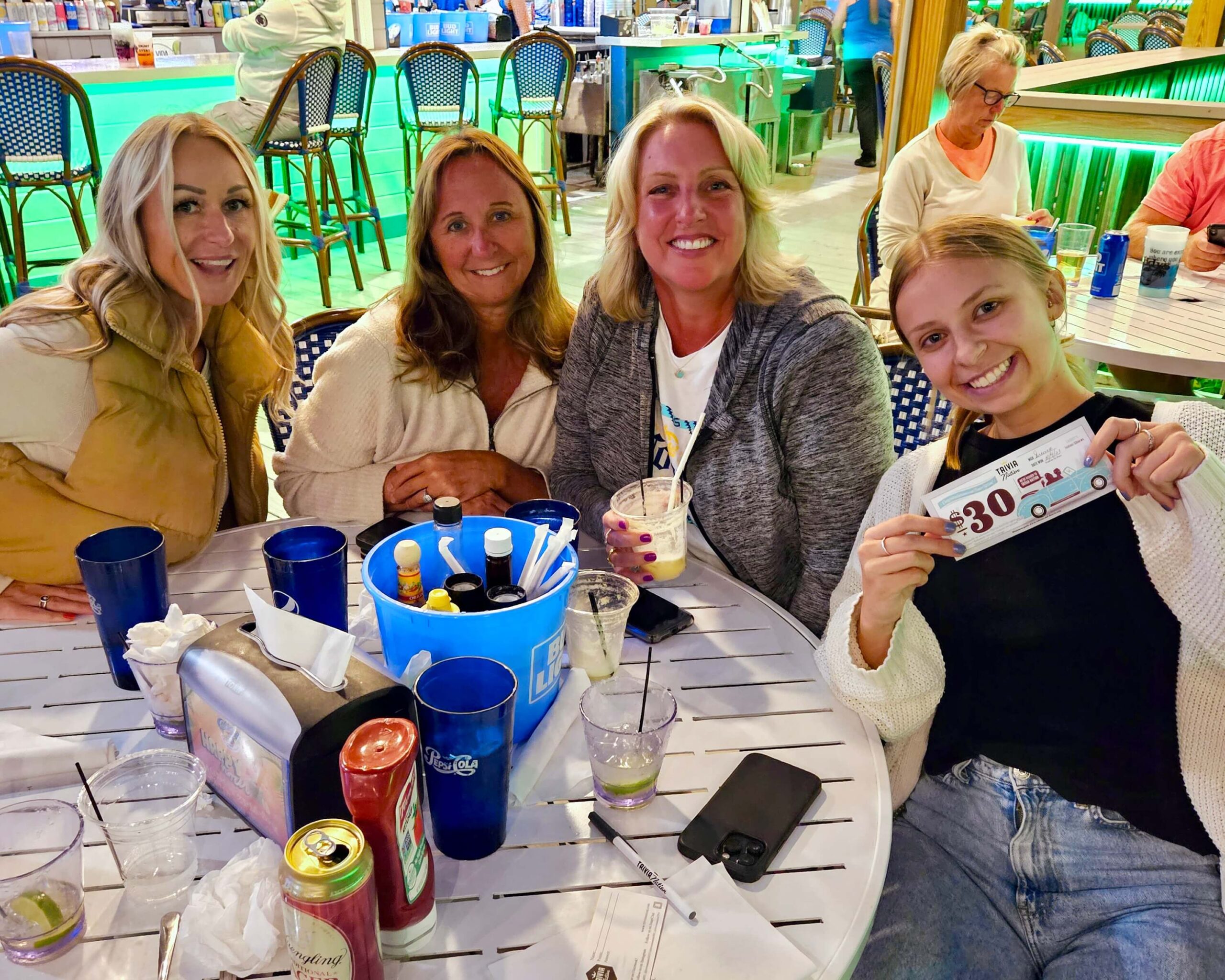 Caddy's Bradenton FL 34208 trivia night: close-up of four women seated around a table on a patio all smiling with drinks on the table and in their hands while the young woman on the right holds up a $30 music bingo coupon