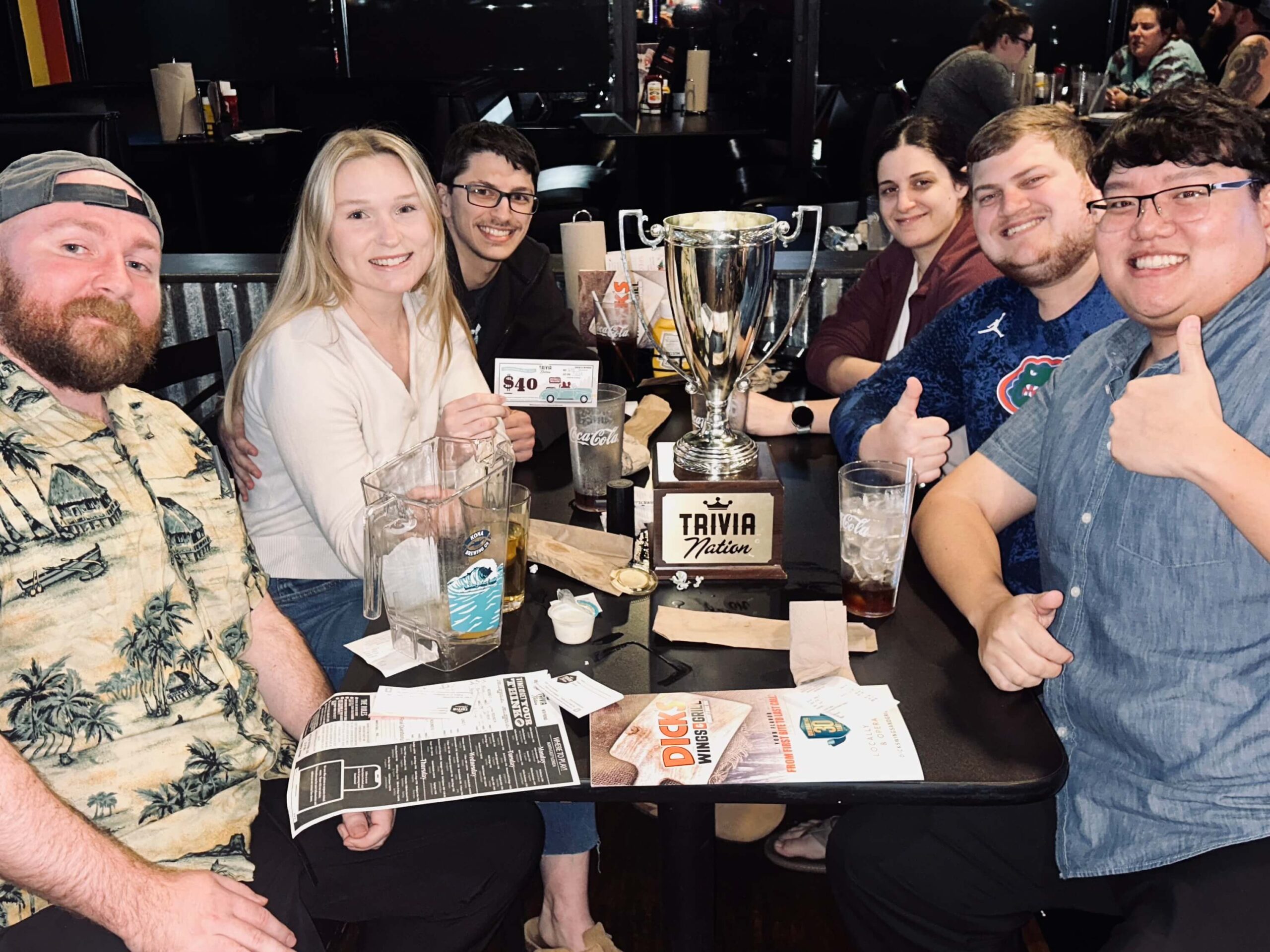 Dick's Wings & Grill Jacksonville FL 32257 trivia night: group of six young adults seated at a table smiling with a Trivia Nation trophy on the table and two of the men holding up "thumbs up" signs