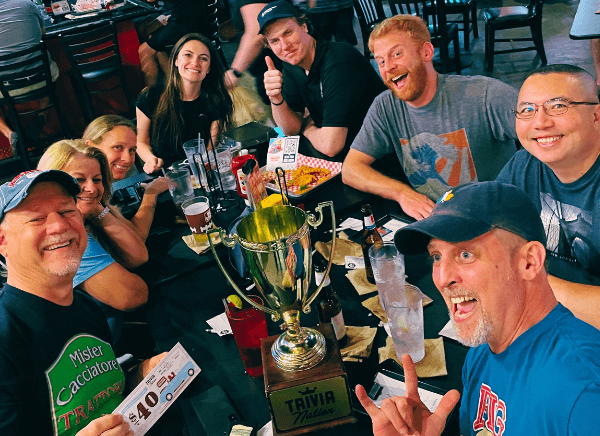 Dick's Wings and Grill Jacksonville FL 32225 trivia night: group of adults both men and women gathered around a table smiling and celebrating with a Trivia Nation trophy on the table and a basket of fries while the man in front makes a funny face and holds up his hand to make a "rock on" sign