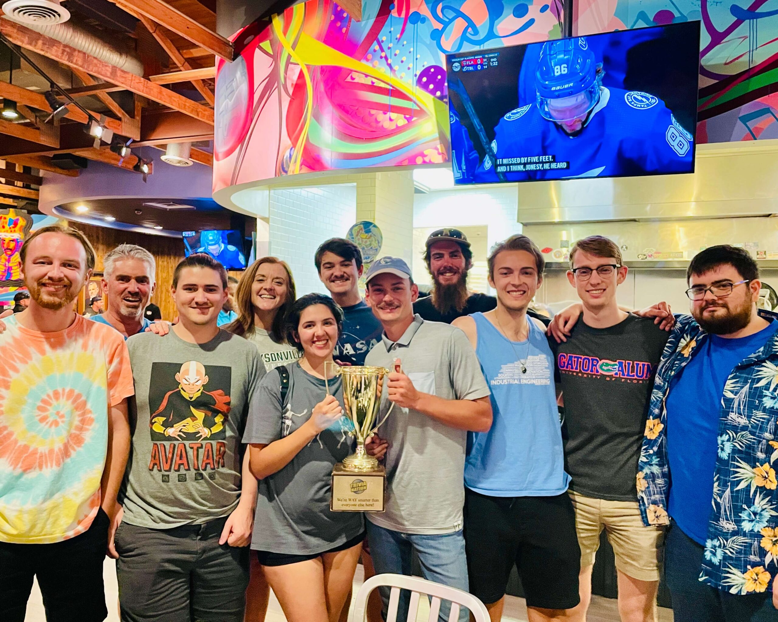 Mellow Mushroom Fleming Island FL 32003 trivia night: large group of young people all standing together smiling while the young woman and man in the middle hold up a Trivia Nation trophy and with a wall above them showing graffiti art and a TV showing hockey