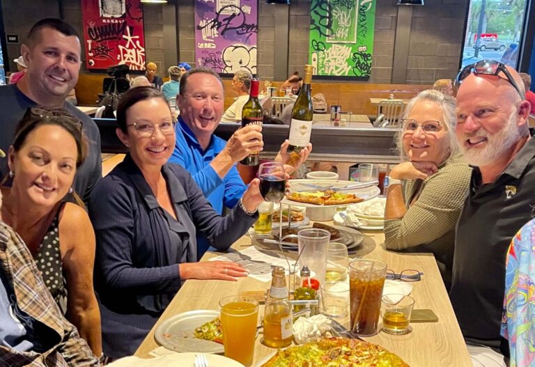 Mellow Mushroom Jacksonville FL 32218 trivia night: group of middle-aged and older adults at a long table smiling with pizza and beers on the table and a man at the end of the table holding up bottles of wine