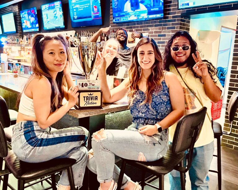 Mellow Mushroom Jacksonville FL 33205 trivia night: two girls seated at a bar smiling with a Trivia Nation trophy between them and a man with long hair next to them smiling
