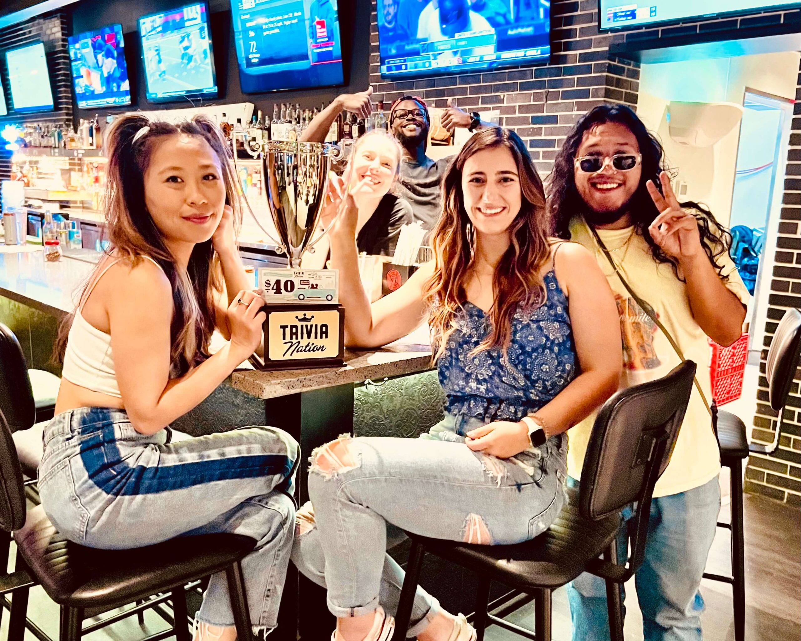 Mellow Mushroom Jacksonville FL 33205 trivia night: two girls seated at a bar smiling with a Trivia Nation trophy between them and a man with long hair next to them smiling