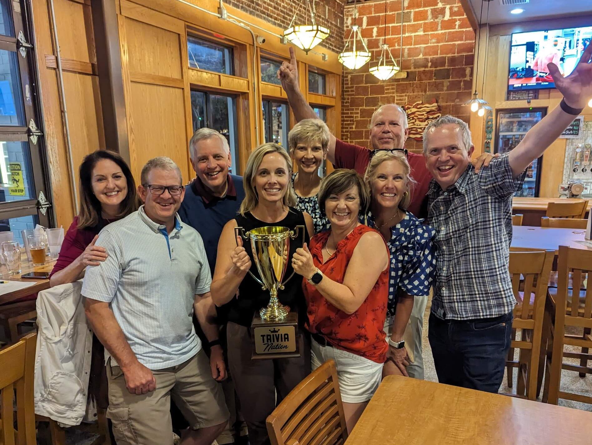 Sanford Brewing Company Sanford FL 32771 trivia night: group of adults smiling and having a good time with a woman in the middle holding a trophy and with bar lights overhead