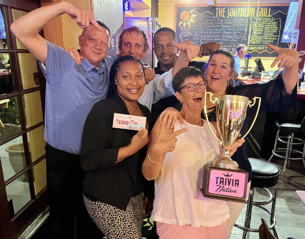 The Southern Grill Jacksonville FL 32207 trivia night: group of adults celebrating together with a woman in front holding a $100 trivia coupon next to another woman holding a Trivia Nation trophy with the other adults behind them pointing at it