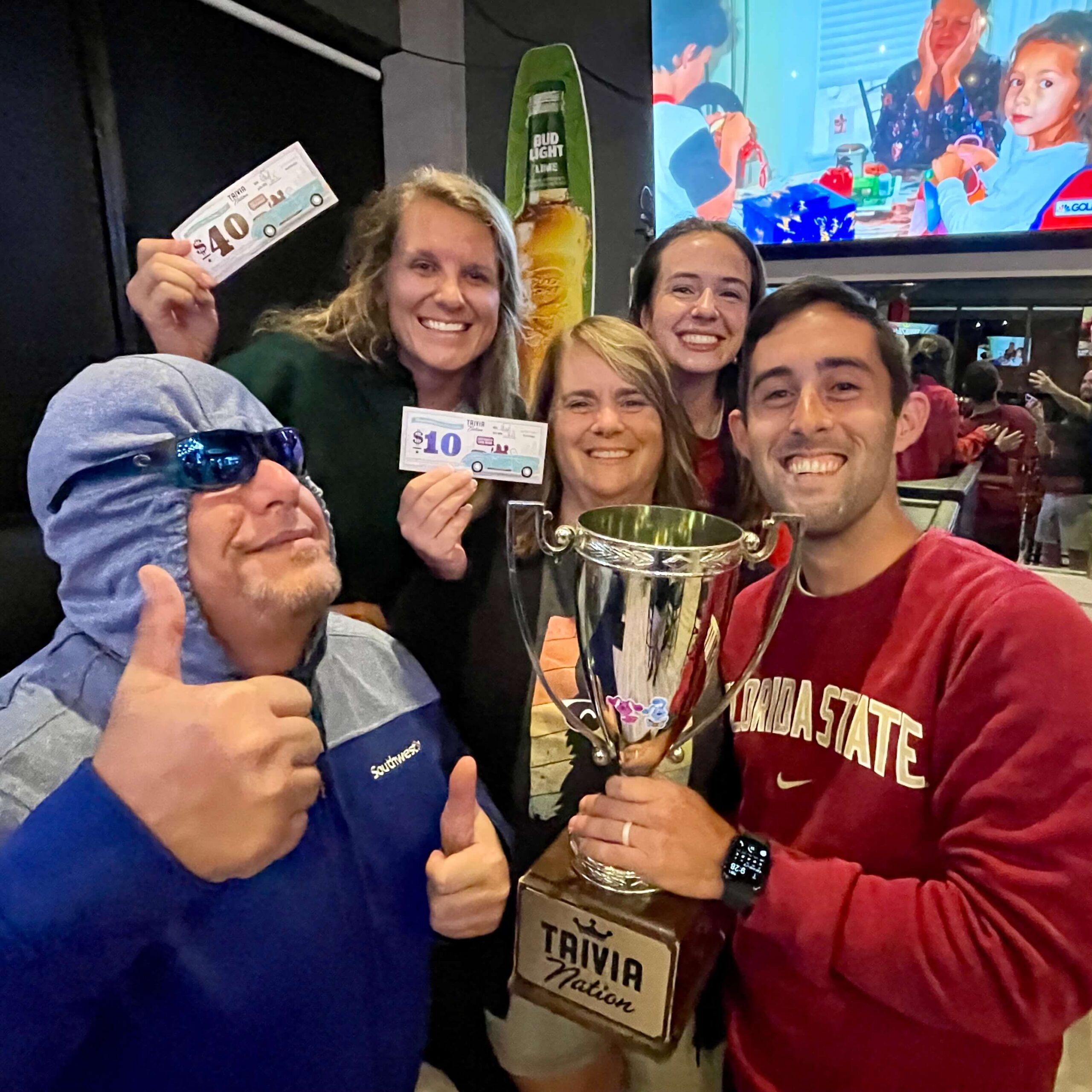 The Twisted Birch Sports Bar & Grill Rockledge FL 32955 trivia night: group of adults having fun with one man having a hoodie tied over his eyes wearing sunglasses and another man smiling and holding up a trophy