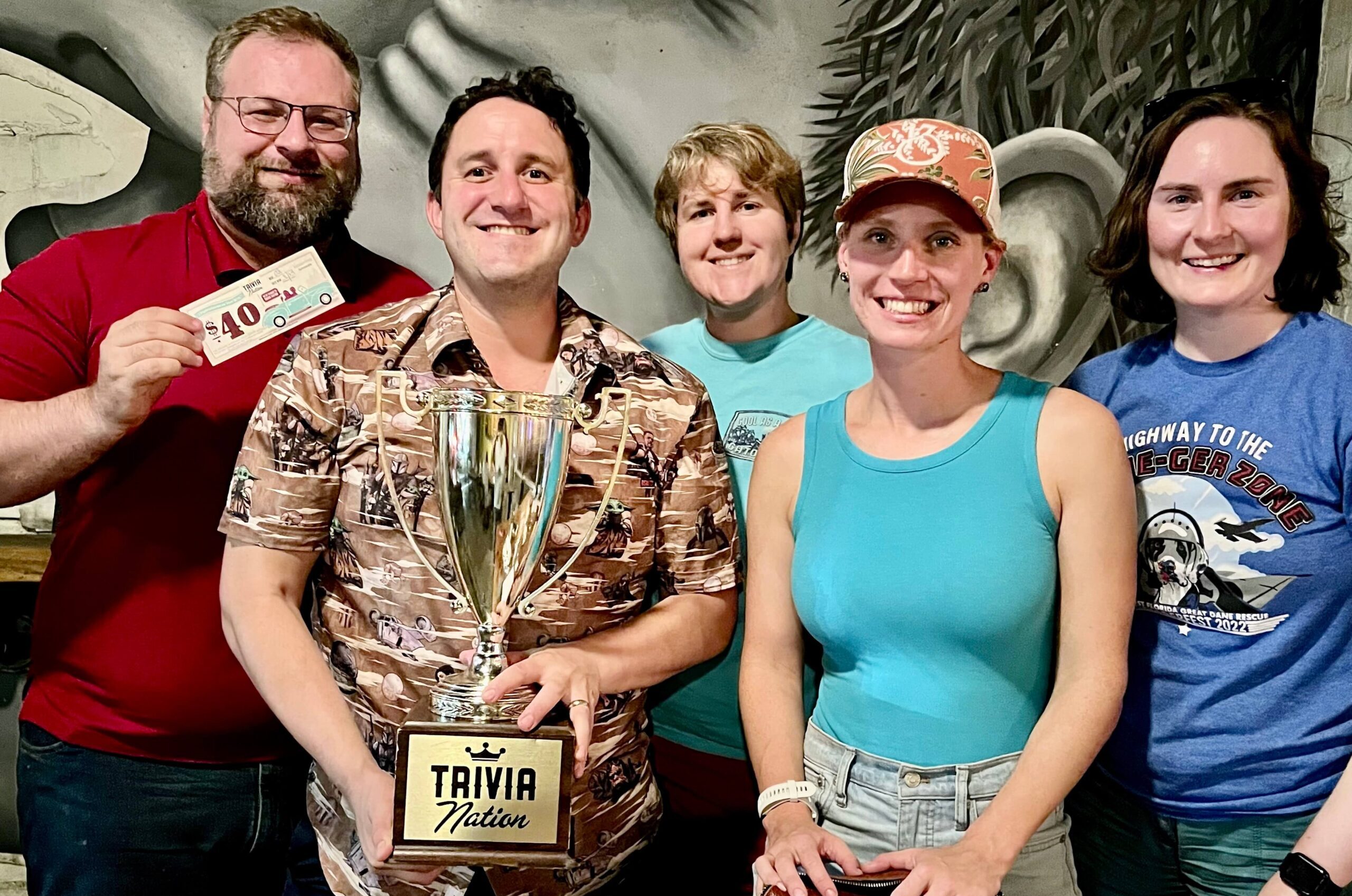 V Pizza & Sidecar Jacksonville FL 32207 trivia night: group of adults in front of a mural wall smiling with a man in front holding up a Trivia Nation trophy and a man in a red shirt behind him holding up a $40 trivia coupon
