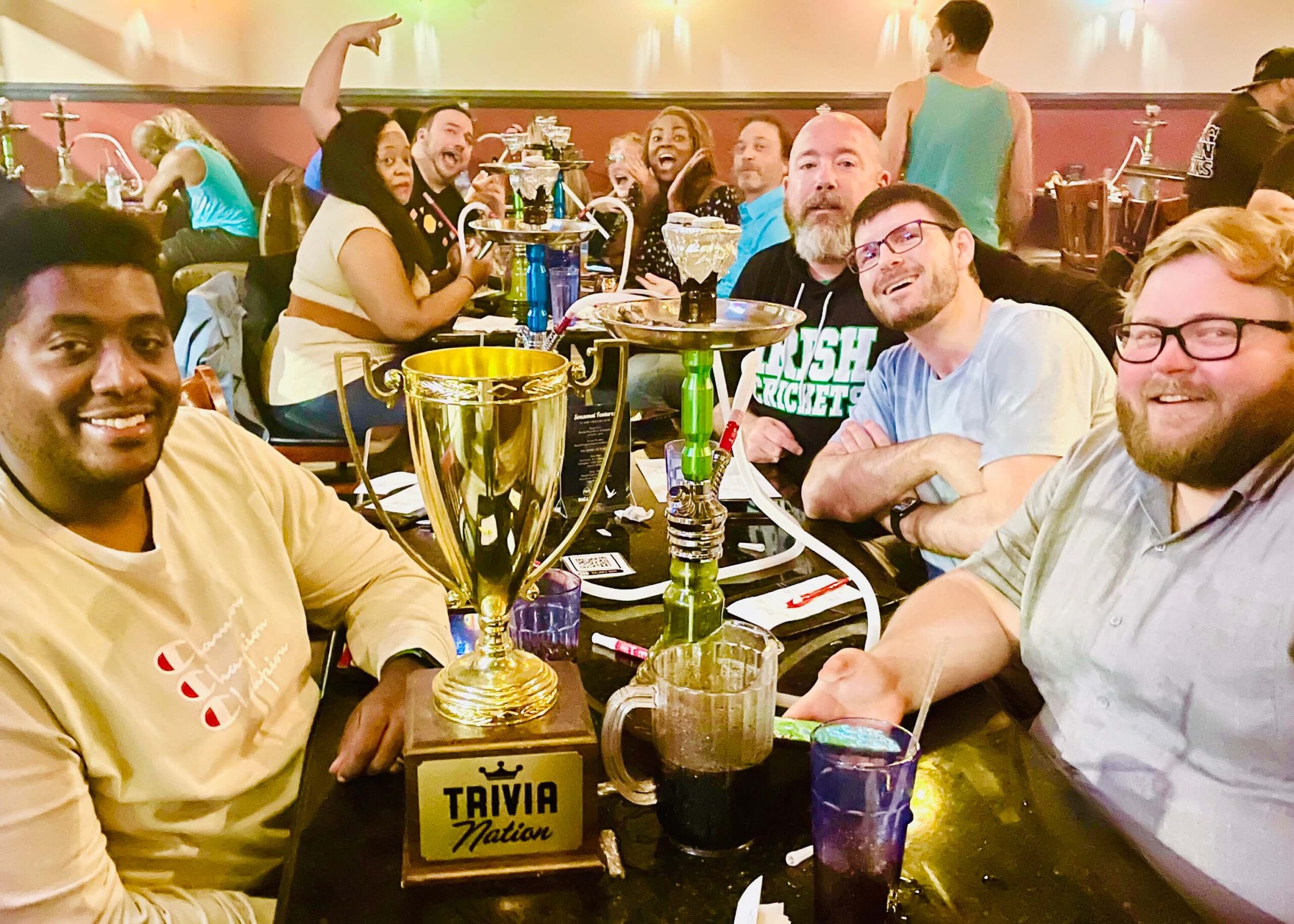 Sahara Cafe & Bar Jacksonville FL 32246 trivia night: table of four men smiling with a Trivia Nation trophy and a hookah with a table full of people also behind them celebrating