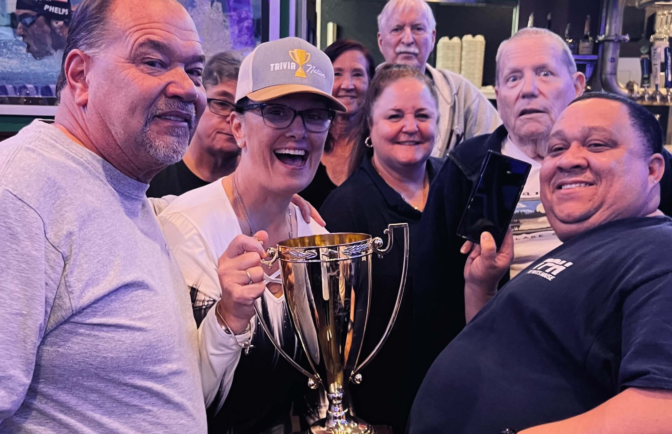 Duffy's Sports Grill Port St. Lucie FL 34593 trivia night: close-up of a group of adults smiling and having fun while the woman in the center wears a Trivia Nation hat and holds up a Trivia Nation trophy