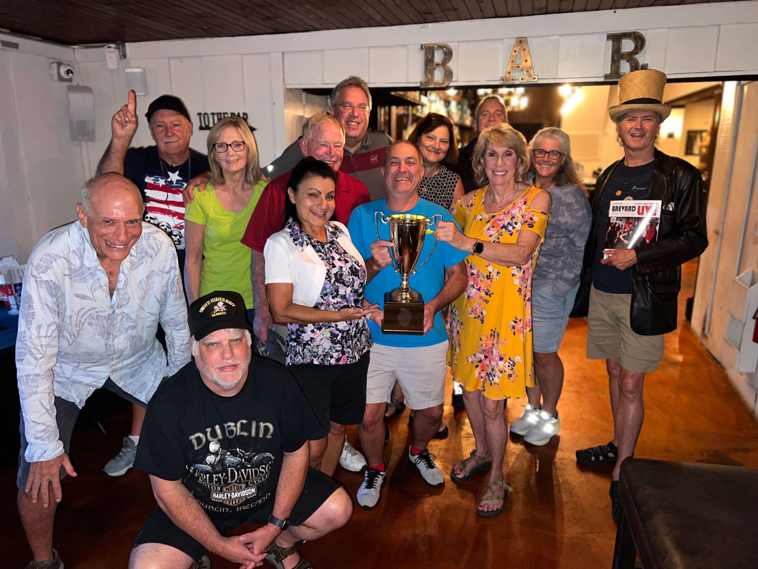 Pineda Inn Bar & Grill Rockledge FL 32955 trivia night: large group of adults smiling and gathered around a trophy in front of a sign that says "BAR" and with one man wearing a top hat