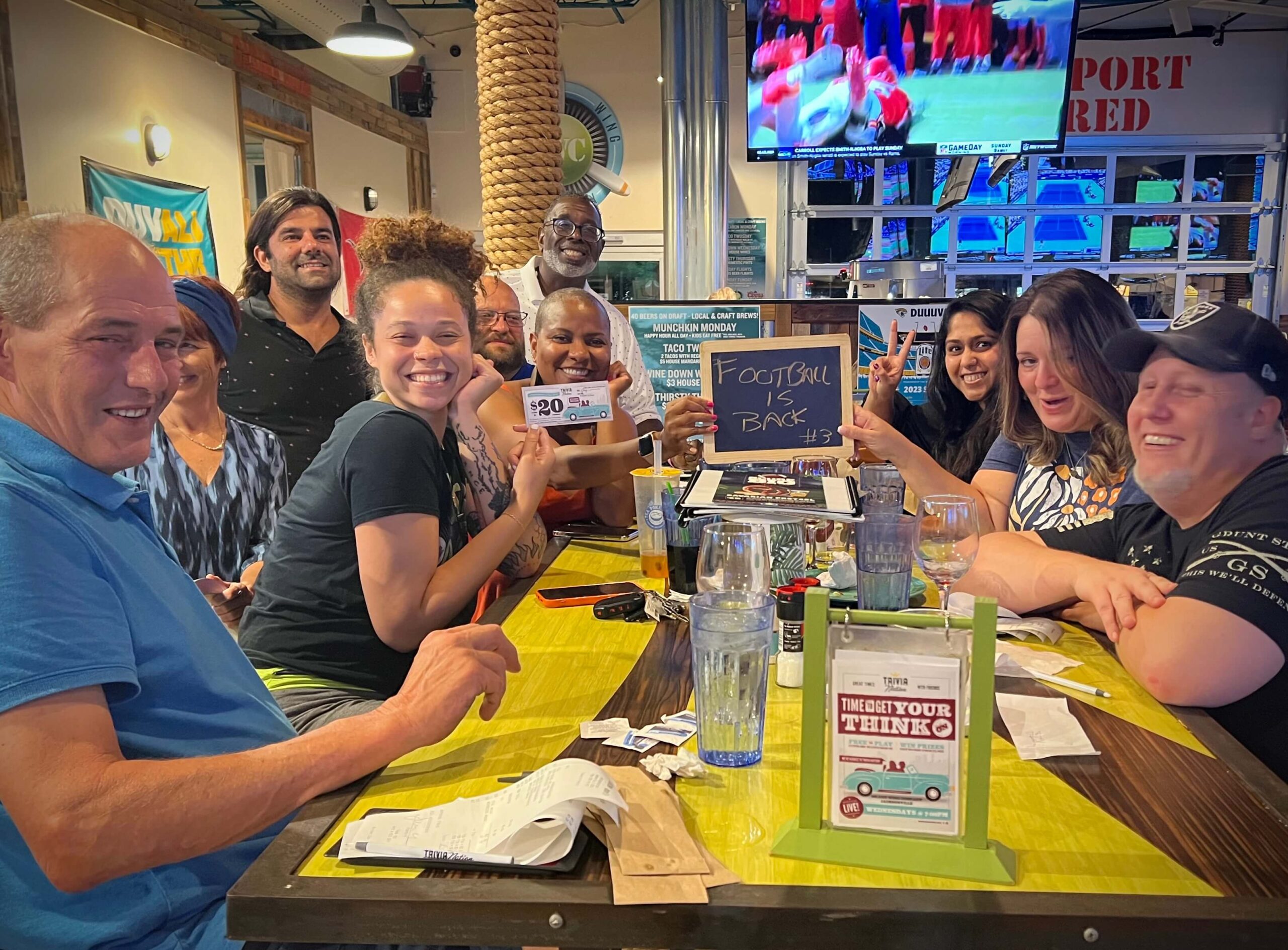 Island Wing Company Jacksonville FL 32216 trivia night: group of adults smiling and laughing while seated around a table and a holding up a chalk board with their trivia team name "Football is Back"