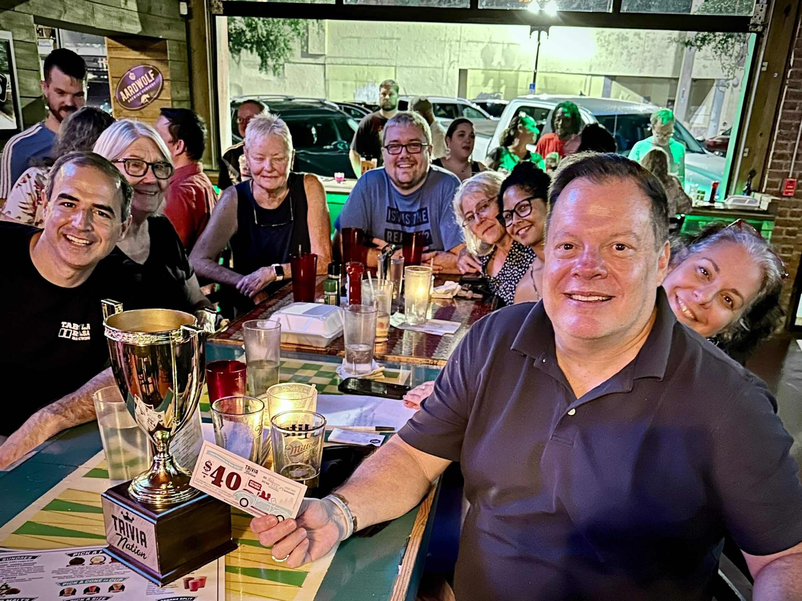 The Stout Snug Jacksonville FL 32205 trivia night: group of adults around a table smiling with an open window overlooking the street behind them and a Trivia Nation trophy on the table