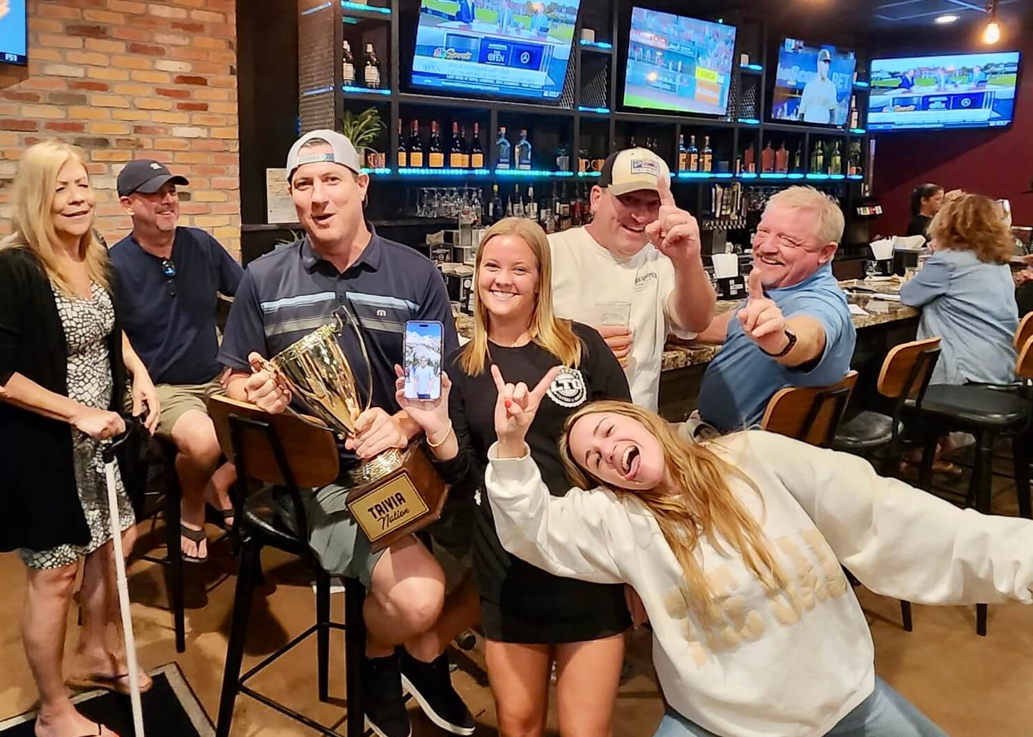 Legends Tavern & Grille Palm Beach Gardens FL 33418 trivia night: group of adults having fun in front of a bar with a man in the middle holding a Trivia Nation trophy and a woman in front making a silly face while two other men hold up their hands making a "number one" sign