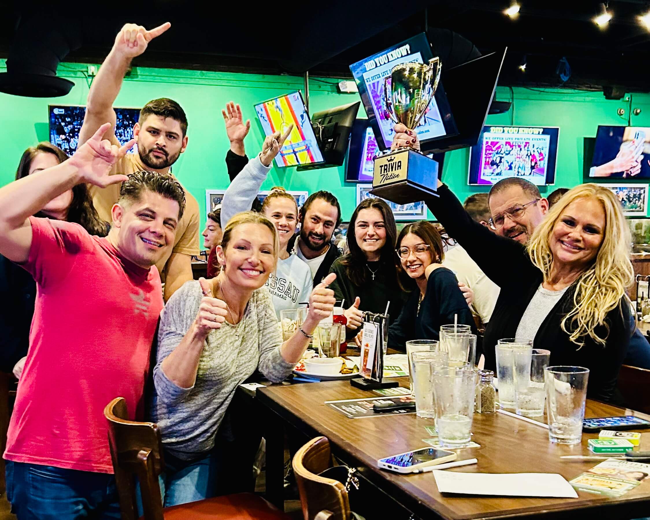 Duffy's Sports Grill Deerfield Beach FL 33441 trivia night: large group of adults having fun and smiling while gathered around a table with some of them holding their hands up and making a "number one" gesture with the woman in front holding a Trivia Nation trophy