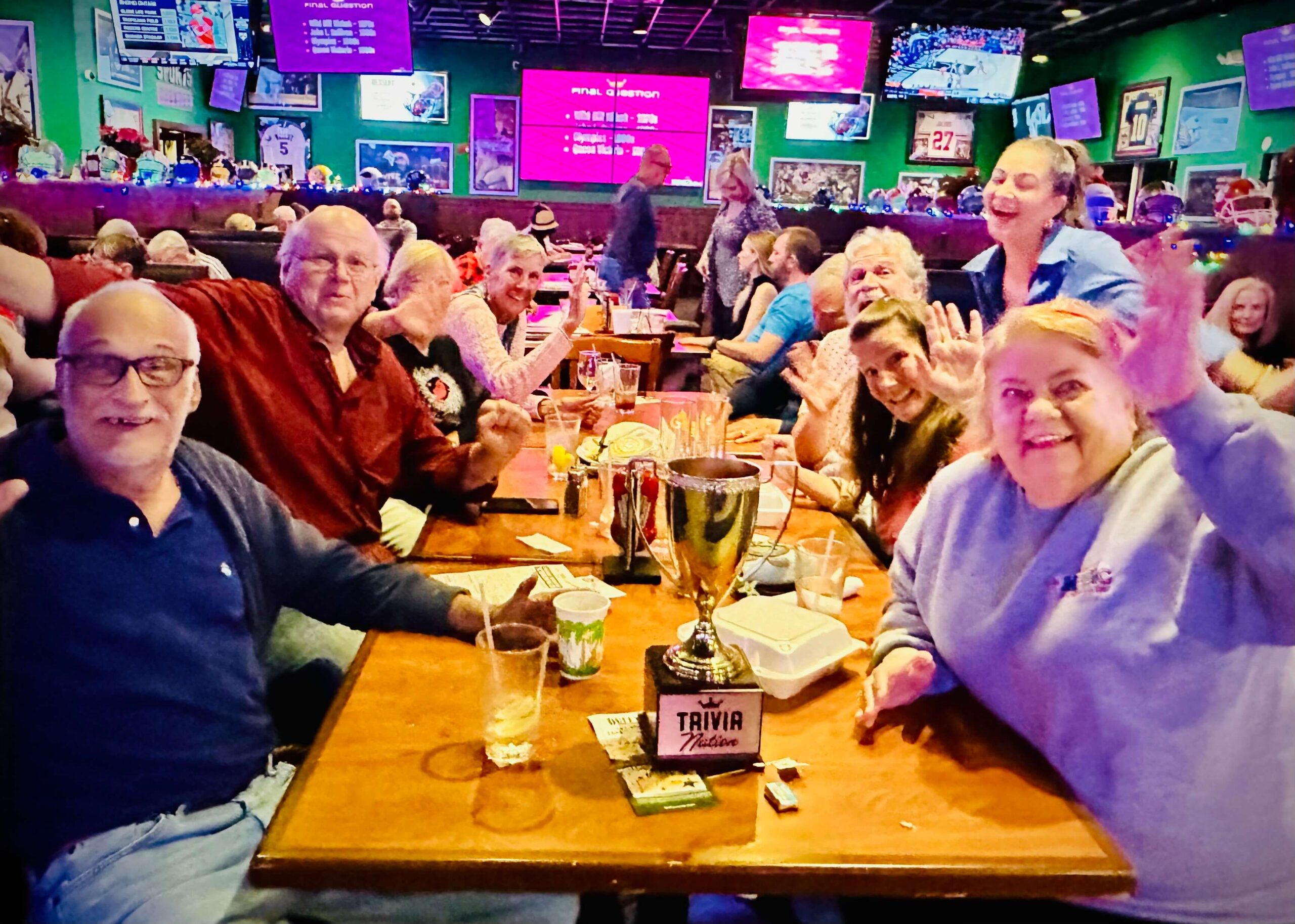 Duffy's Sports Grill Port St. Lucie FL 34986 trivia night: group of adults gathered around a table having fun and raising their hands up with a Trivia Nation trophy on the table and tables of other restaurant patrons behind them