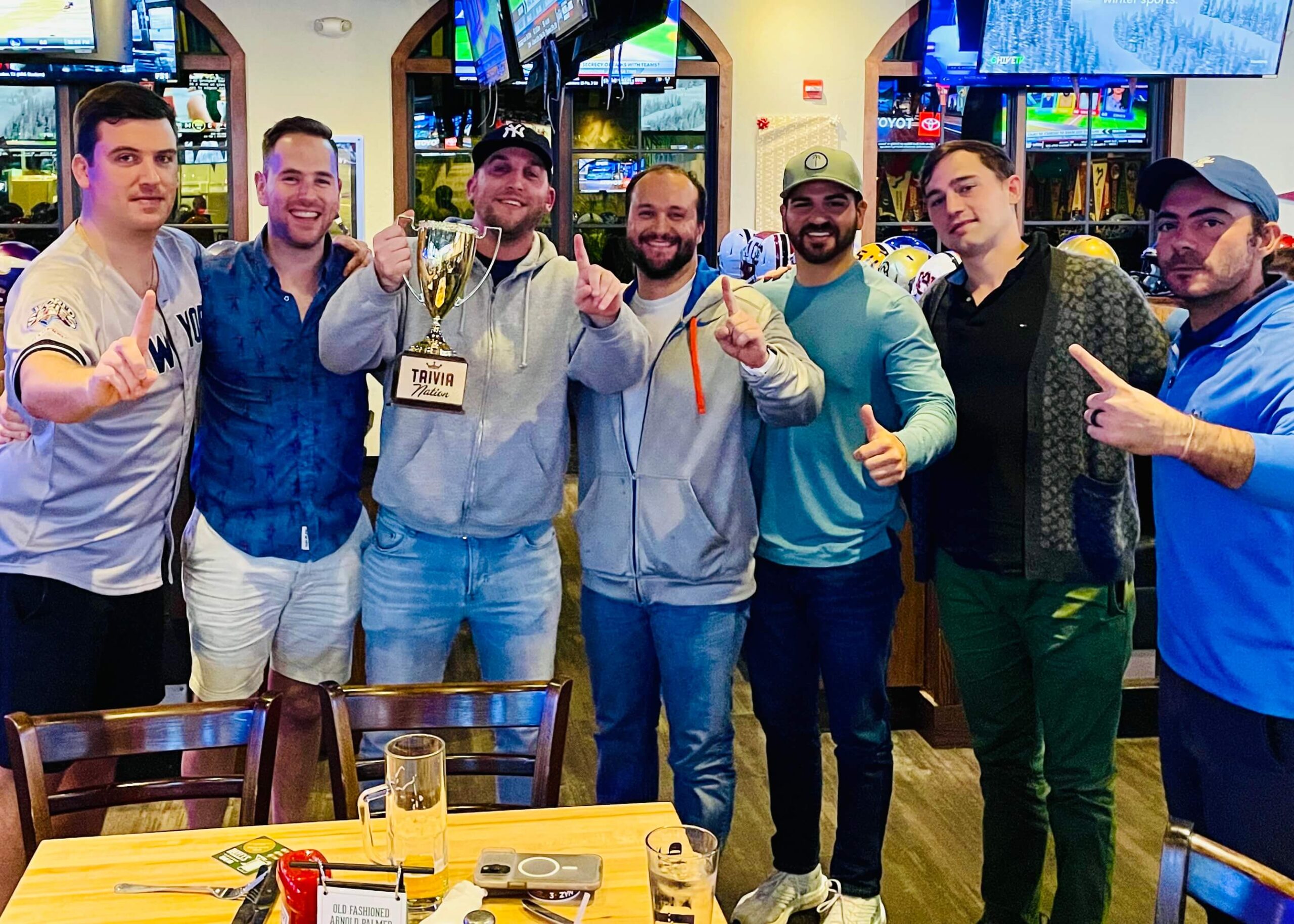 Duffy's Sports Grill Tampa FL 33607 trivia night: group of seven men standing together in a line all smiling and holding up their hands in a number one gesture with one man in the middle holding up a Trivia Nation trophy