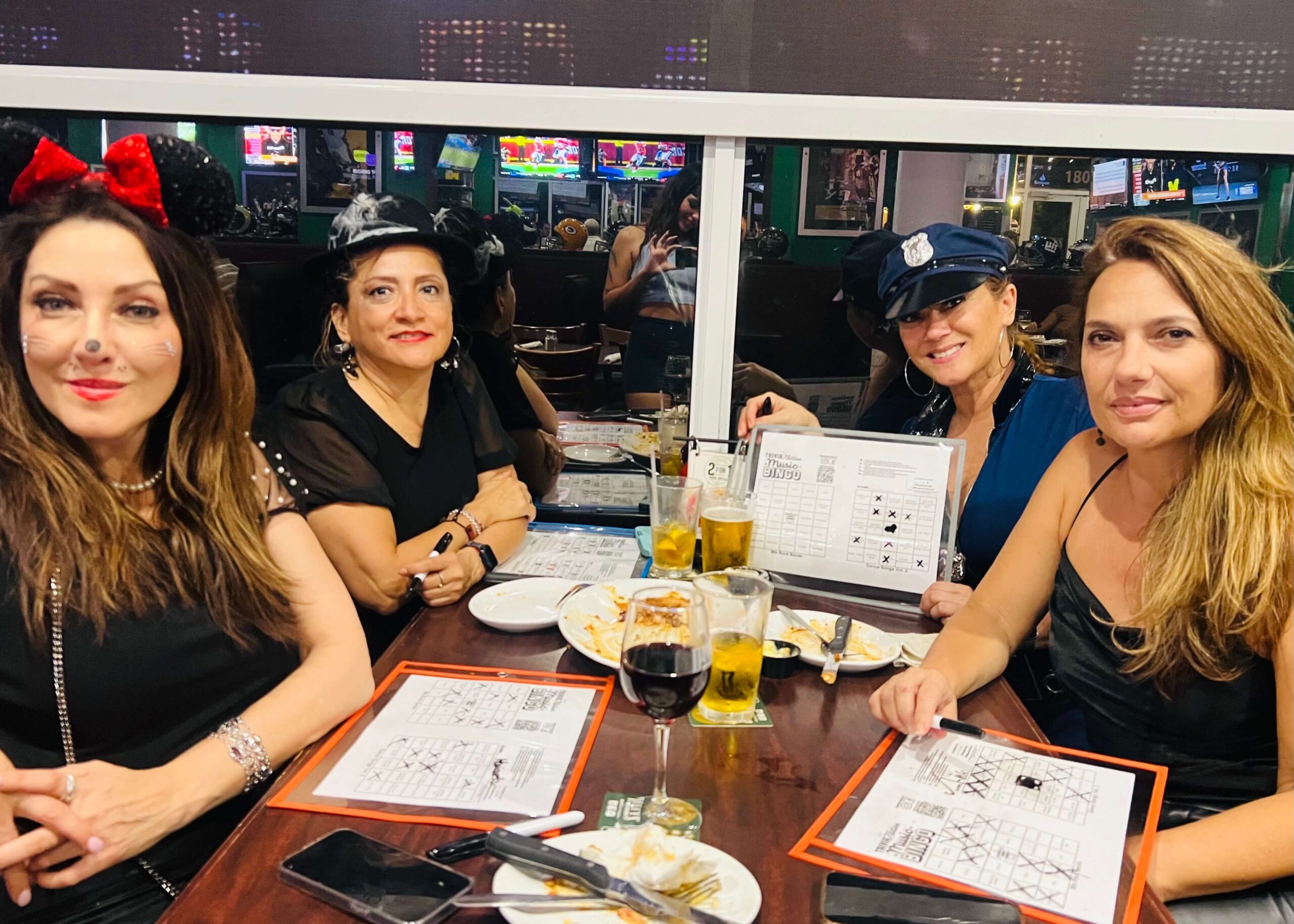 Duffy's Sports Grill Weston FL 33326 trivia night: group of four middle-aged attractive women seated at a table with music bingo cards and beer and wine on the table with three of the women wearing costumes for Halloween