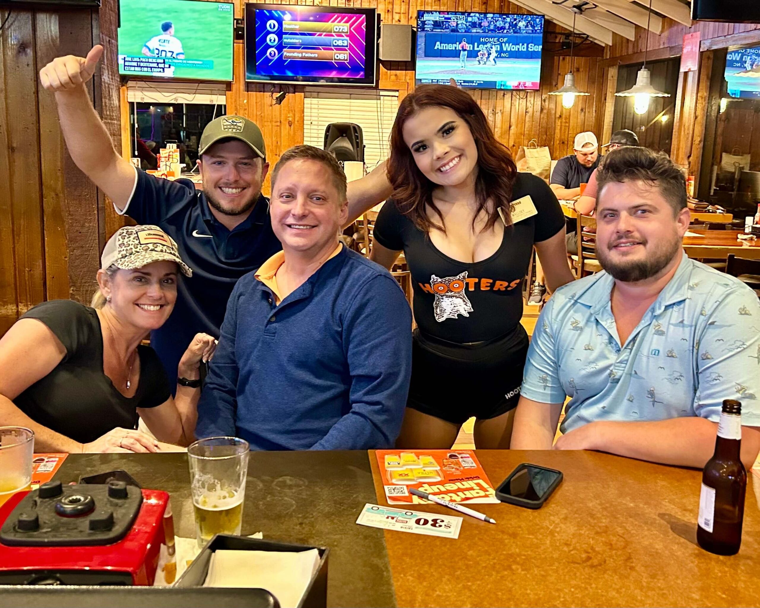 Hooters Jacksonville FL trivia night 32257: five adults at a bar smiling including a Hooters waitress with two draft beers and a bottle of beer on the bar