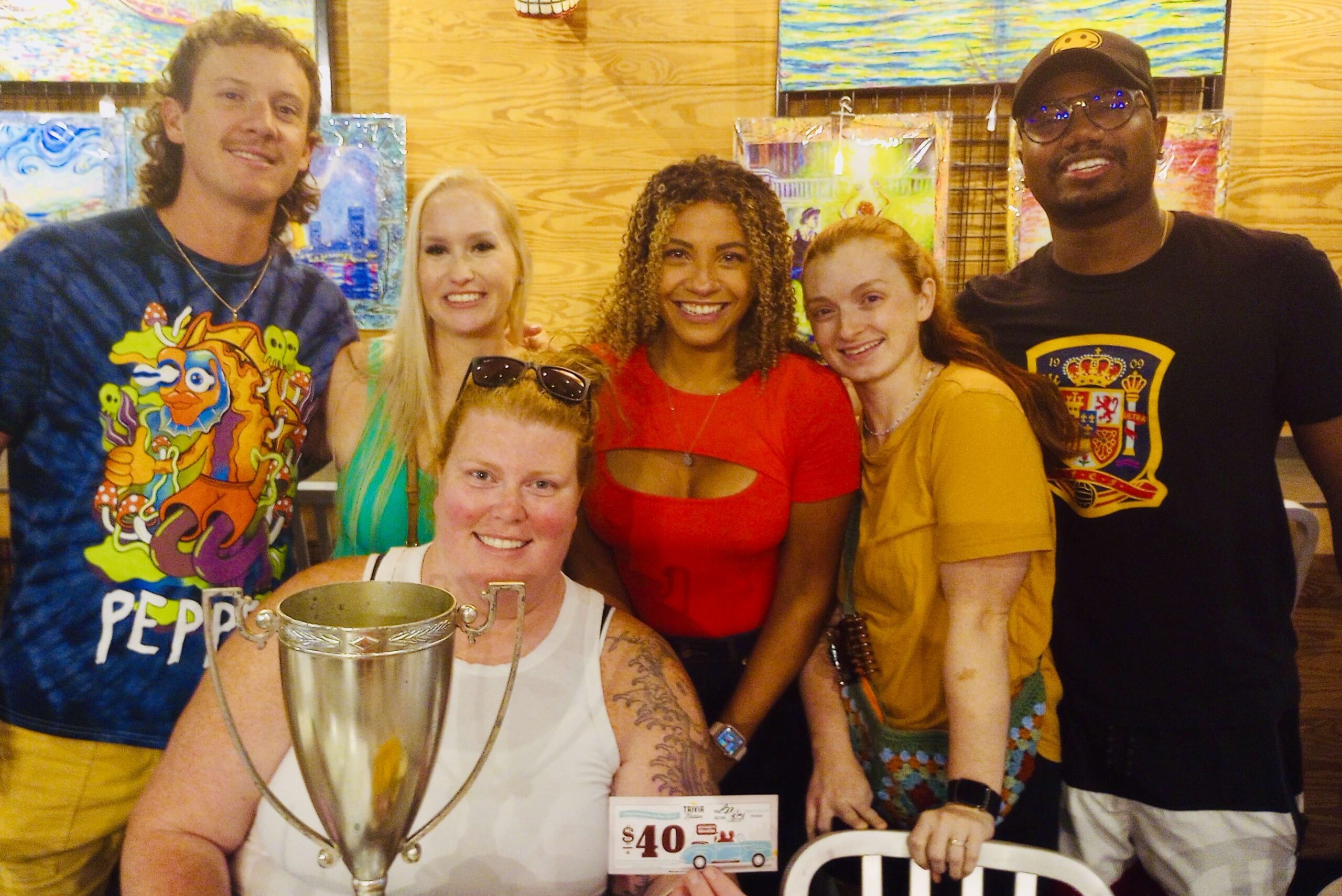Burrito Gallery Jacksonville FL 32202 trivia night: group of diverse young people both men and women standing together smiling with the woman in front seated behind a trophy and holding up a $40 trivia coupon