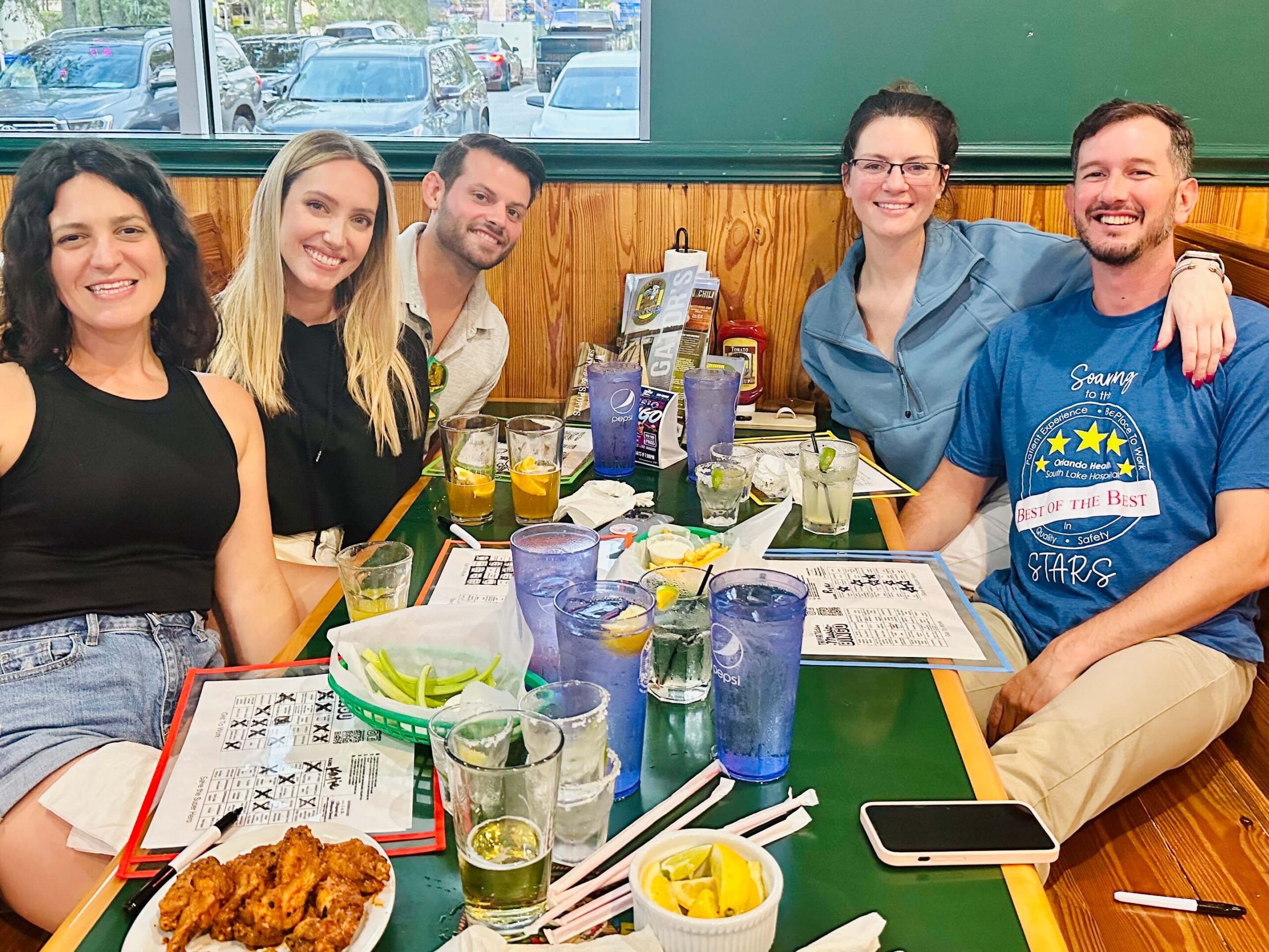 Gators Dockside Jacksonville FL 32222 trivia night: group of attractive young men and women seated around a green table with food including wings on the table as well as music bingo cards
