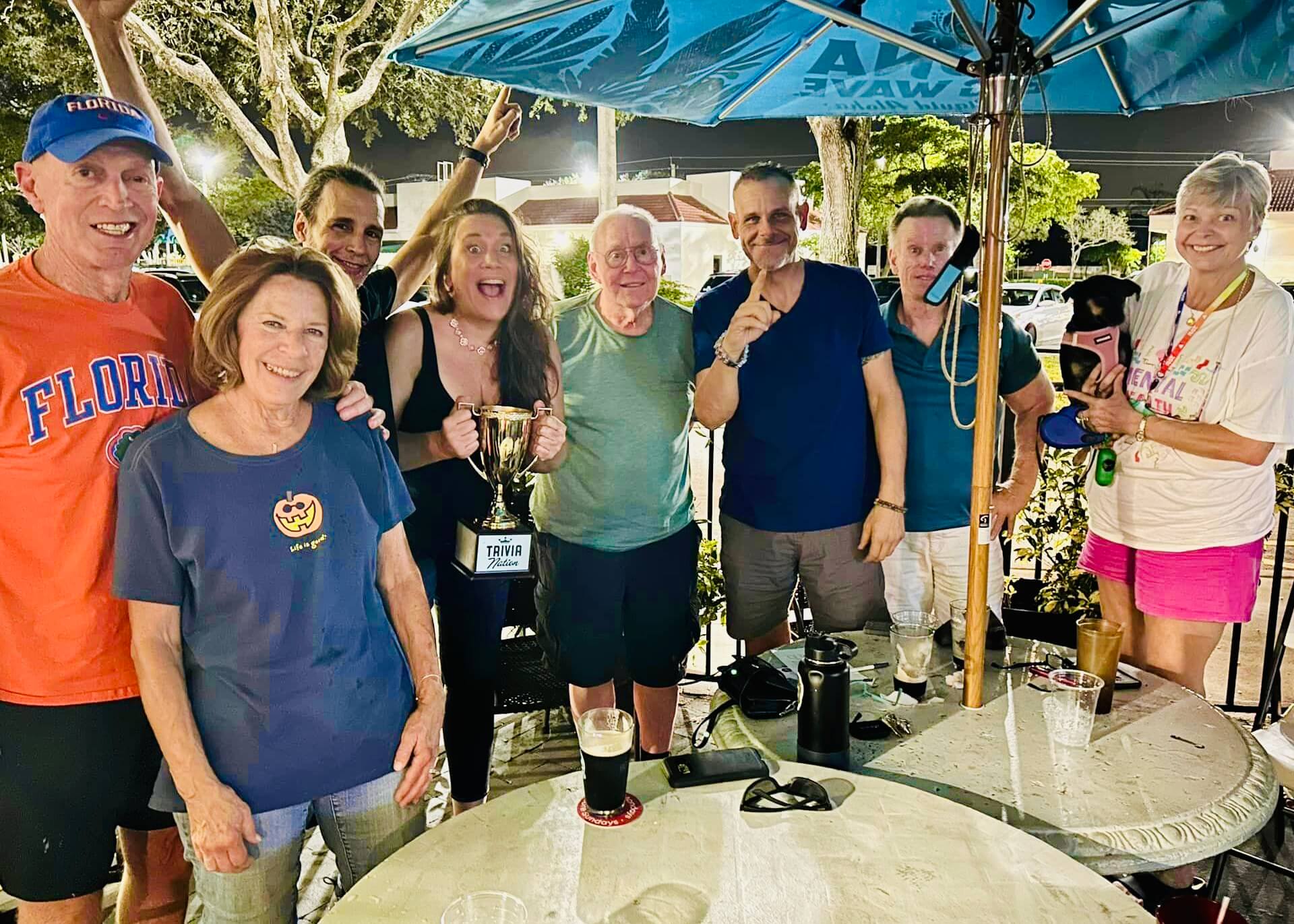 Sharkey's Bar & Grill Coral Springs FL 33065 trivia night: group of adults from middle-aged to older standing outside together smiling with a woman making an excited face and holding a Trivia Nation trophy while another woman holds up a dog