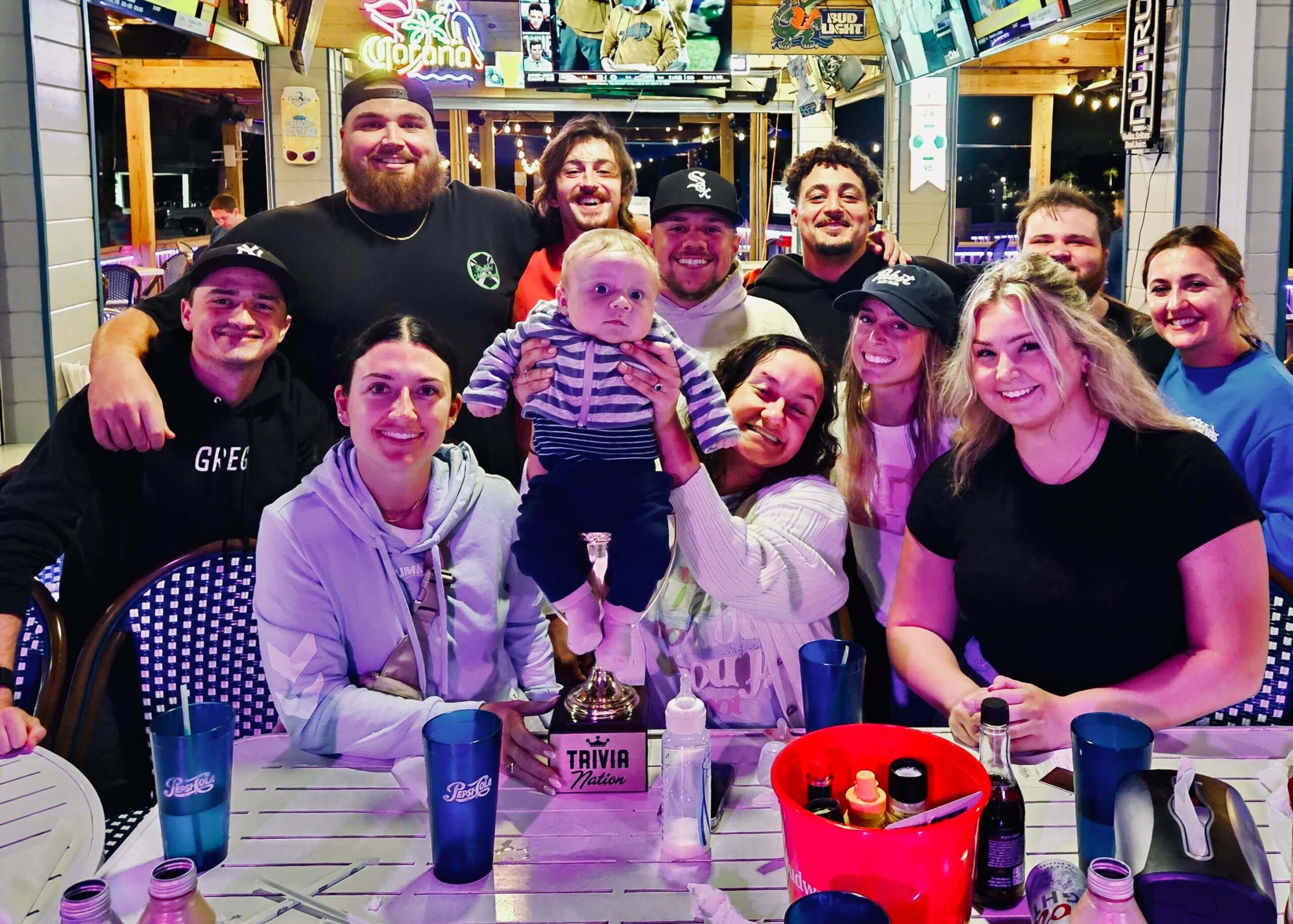 Caddy's Indian Shores FL 33785 trivia night: group of young people all standing together in front of a table smiling while a woman in front holds up a baby seated on top of a Trivia Nation trophy