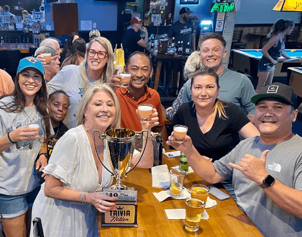 Cliff's Bar & Grill Jacksonville FL 32225 trivia night: group of diverse adults having fun and gathered around a table holding up shot glasses in their hands with beers on the table and a Trivia Nation trophy