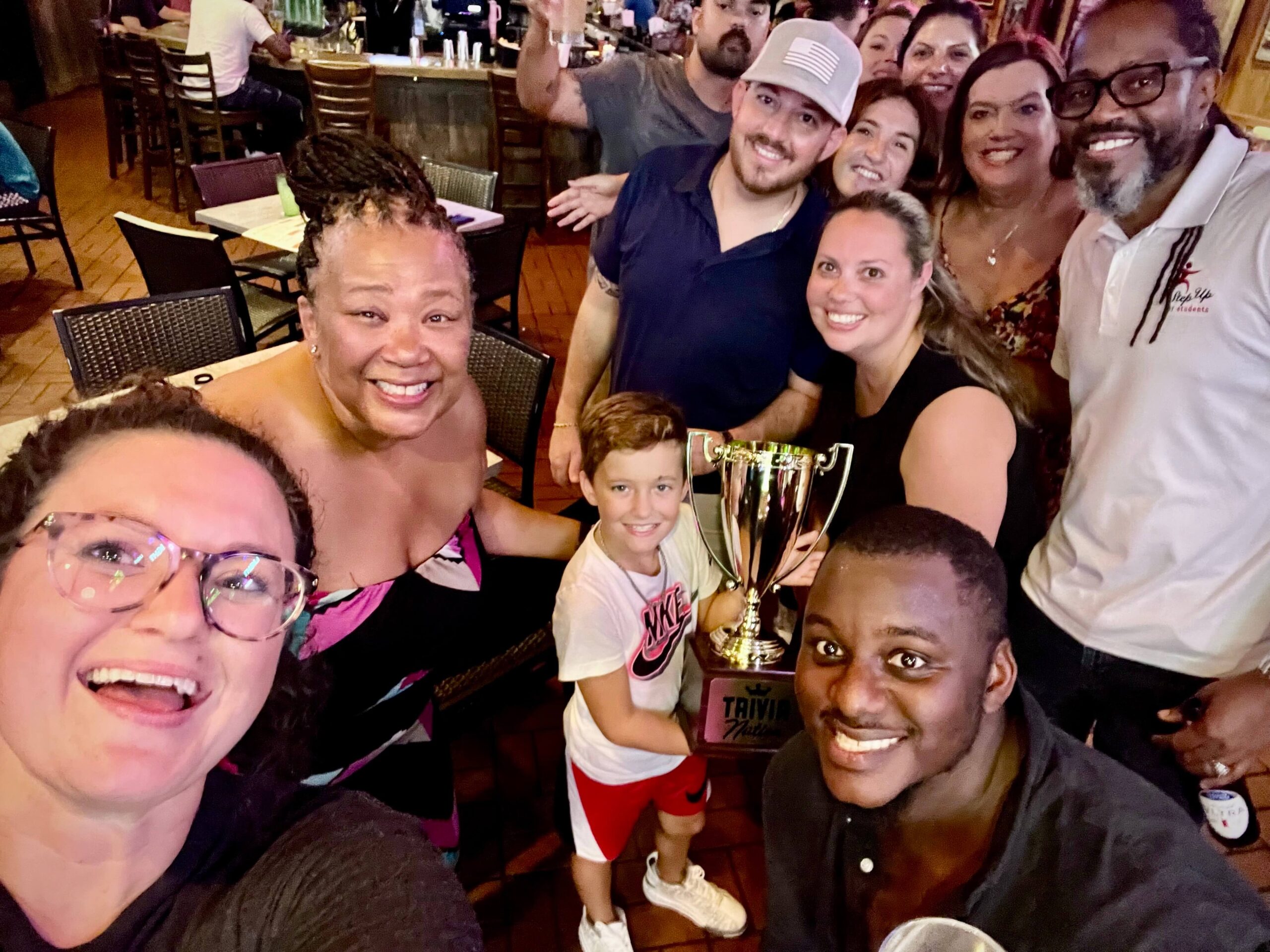 Miller's Ale House Jacksonville FL 32246 trivia night: a large group of people all smiling and leaning in together with a young boy in the middle holding up a Trivia Nation trophy