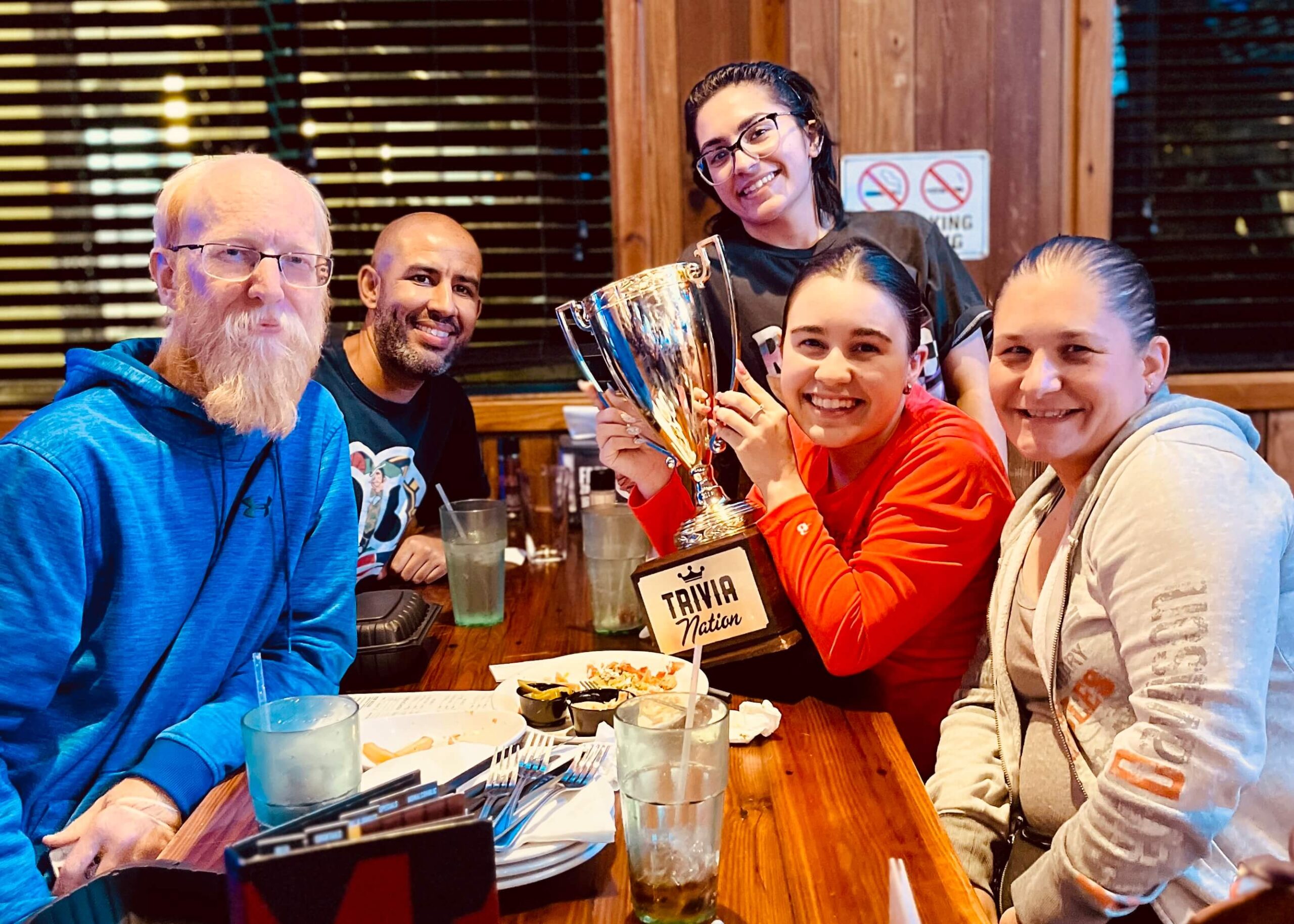 Miller's Ale House Miami FL 33176 trivia night: group of five men and women sitting at a table smiling with empty plates of food on the table and a woman in an orange shirt holding a Trivia Nation trophy