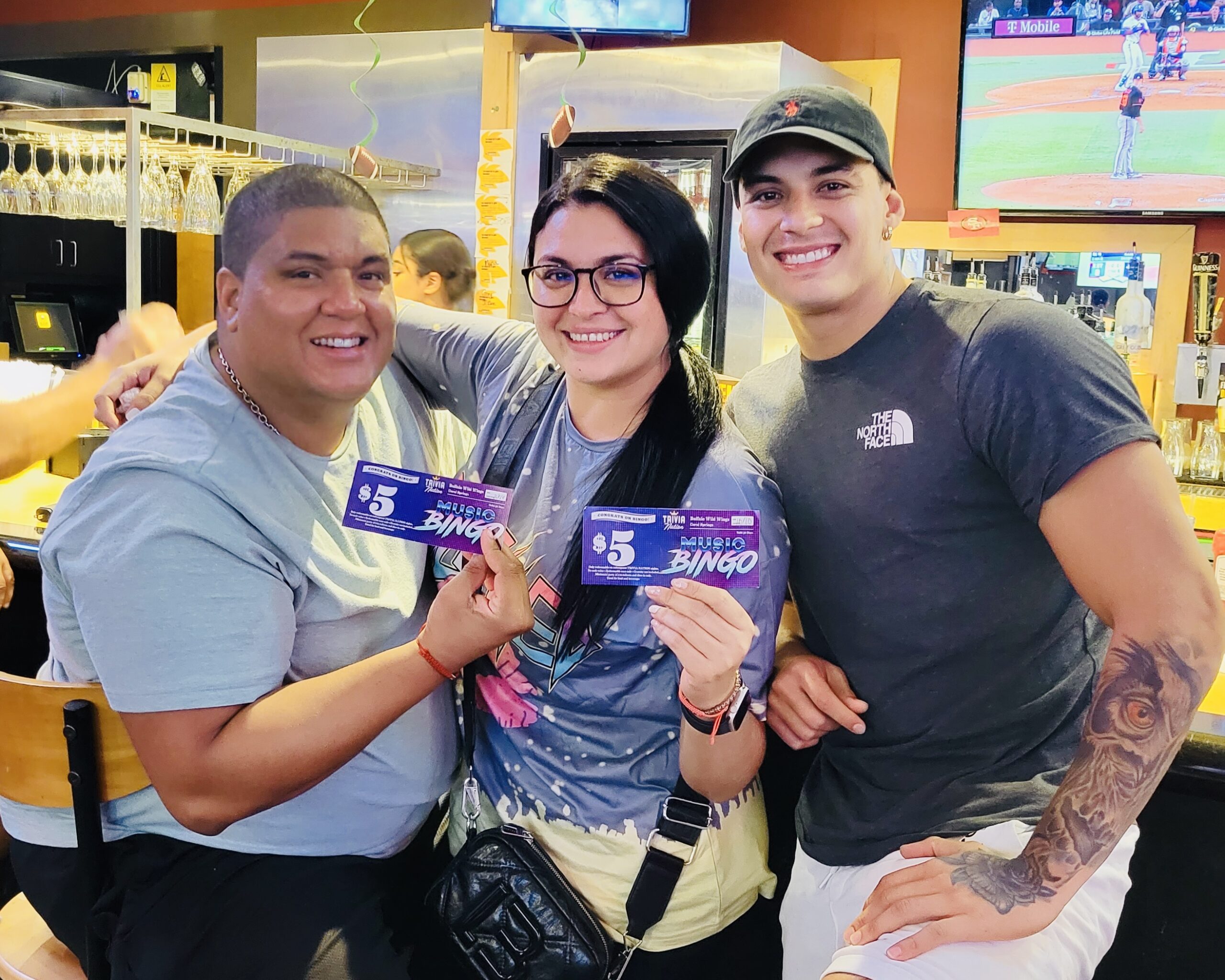 Buffalo Wild Wings Coral Springs FL 33067 trivia night: three adults, two men and one woman, standing together holding up music bingo coupons and smiling with a bar in the background