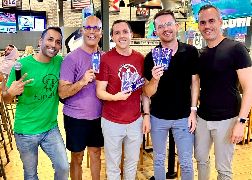 Buffalo Wild Wings Deerfield Beach FL 33441 trivia night: group of five men standing together in a group smiling with the three men in the middle holding up music bingo cards and the male host on the left holding a microphone