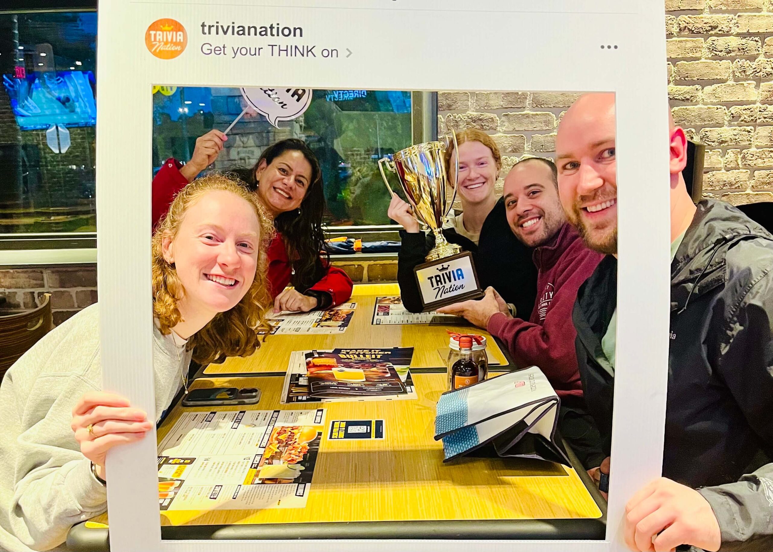 Buffalo Wild Wings Plantation FL 33388 trivia night: group of young adults and a woman gathered together around a table smiling with Trivia Nation props and a trophy including an Instagram Polaroid framing their faces