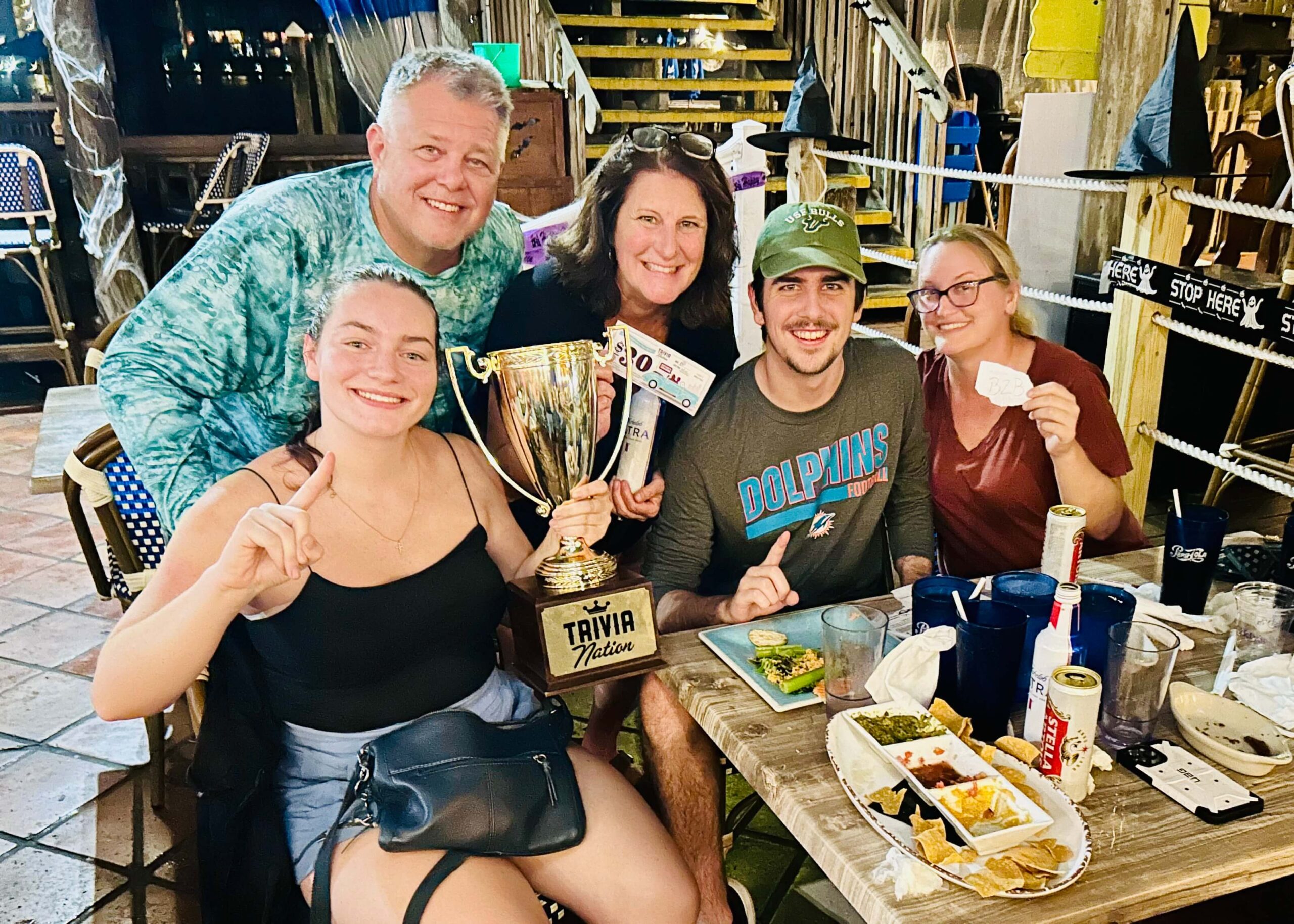 Caddy's John's Pass Madeira Beach FL 33708 trivia night: close-up of a family gathered together at a table smiling around a Trivia Nation trophy and plates of food and drinks