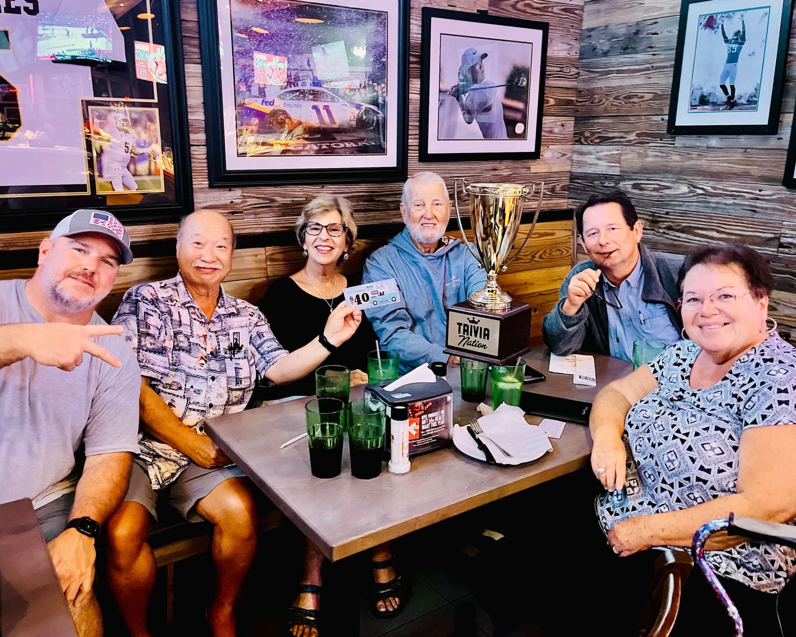 Miller's Ale House Daytona Beach FL 32117 trivia night: group of adults seated around a table all smiling with the trivia host while one man in the back holds up a Trivia Nation trophy and with framed photos of sports scenes on the wall behind them