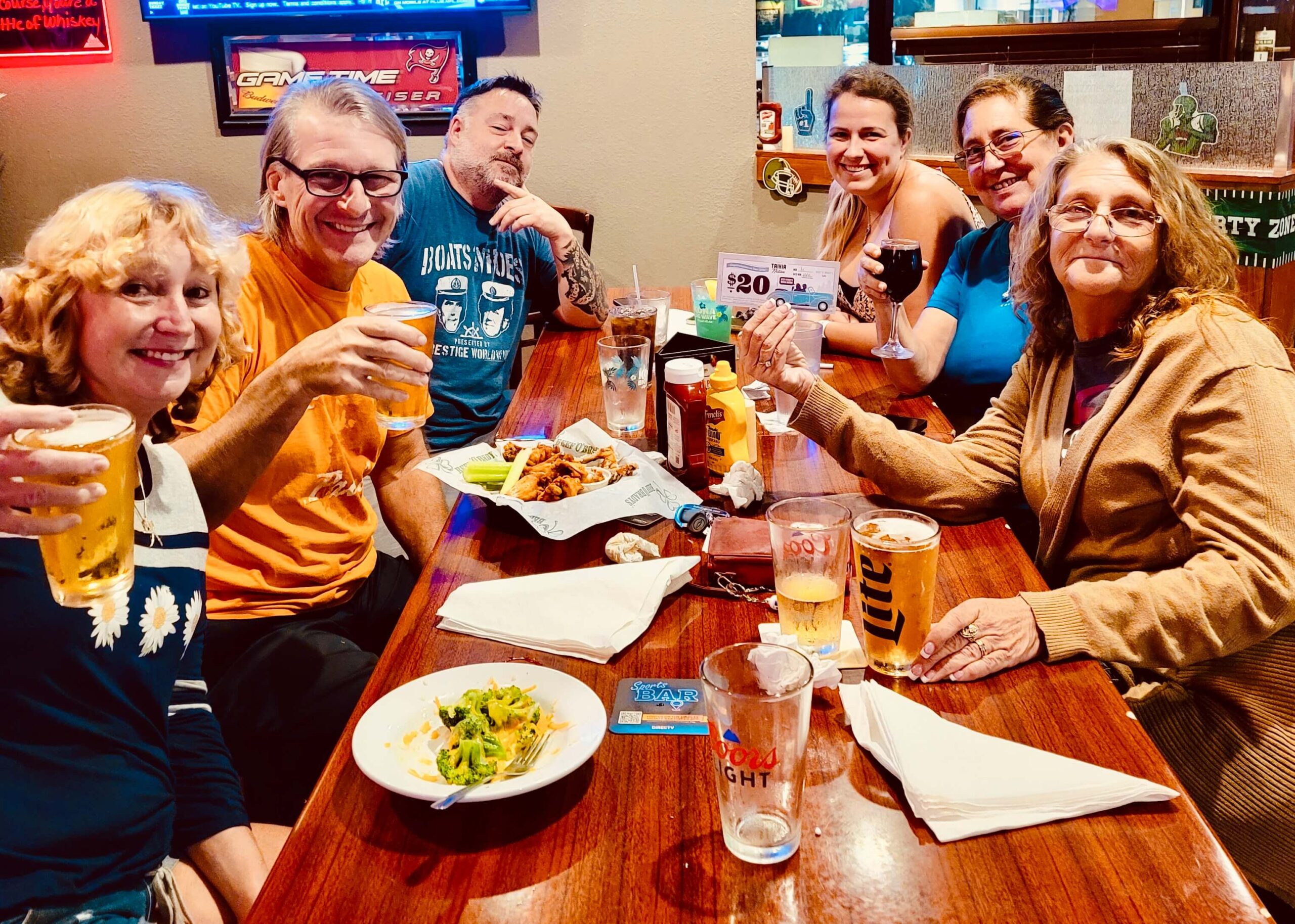 Beef 'O' Brady's Bartow FL 33830 trivia night: group of six adults gathered around a table smiling with wings and broccoli on the table while they hold up glasses of beer, wine, and a $20 trivia coupon
