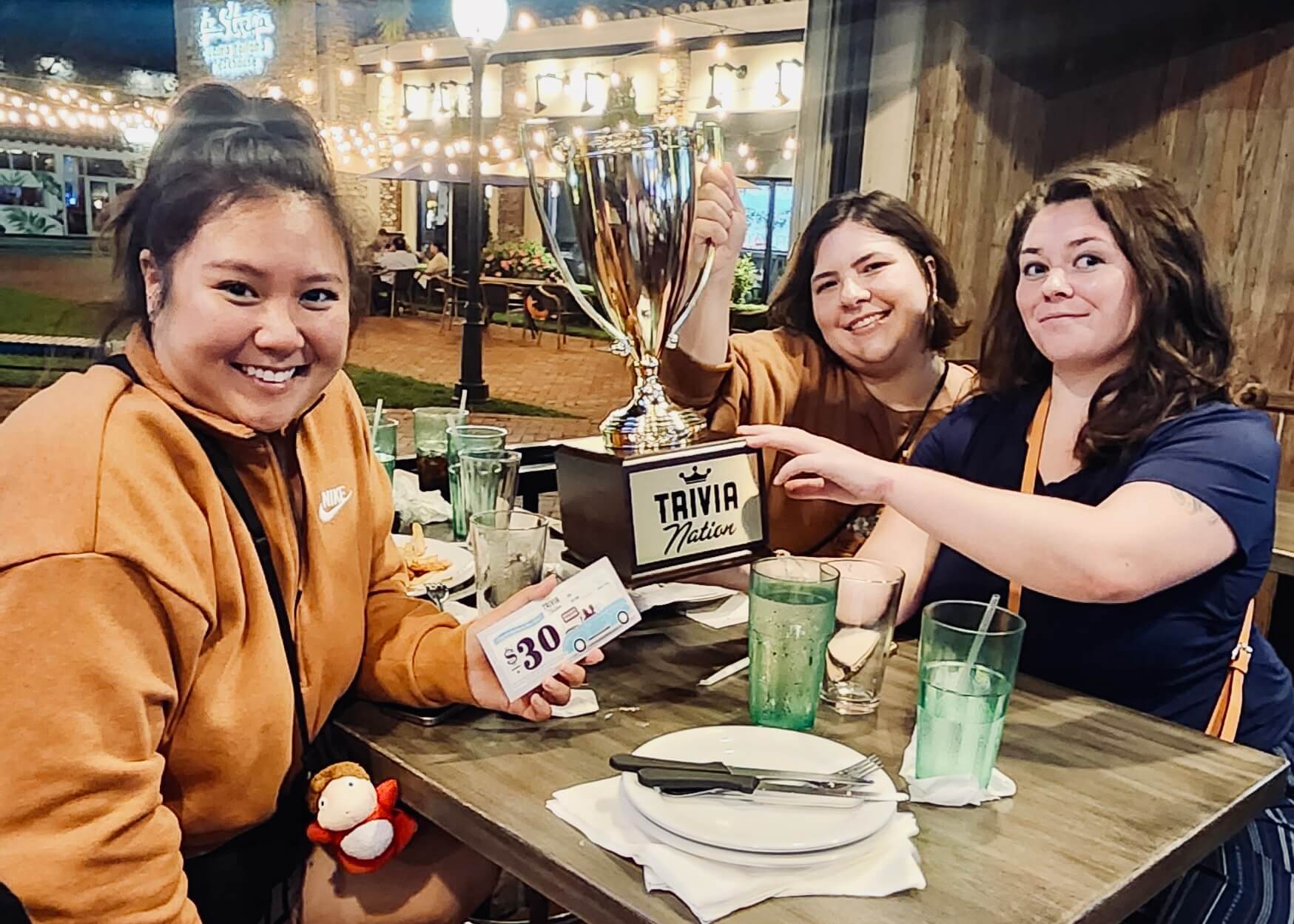 Miller's Ale House Miami Lakes FL 33014 trivia night: three young women seated at a table smiling while holding up a Trivia Nation trophy and coupon with a lit courtyard behind them in the background