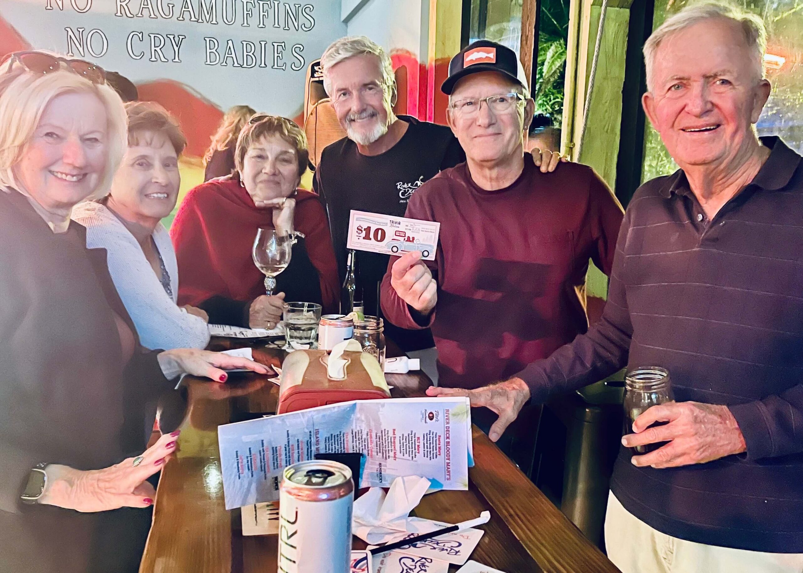 River Deck Tiki Bar & Restaurant New Smyrna Beach FL 32168 trivia night: group of older men and women smiling and standing around a table with one man holding up a $10 trivia coupon